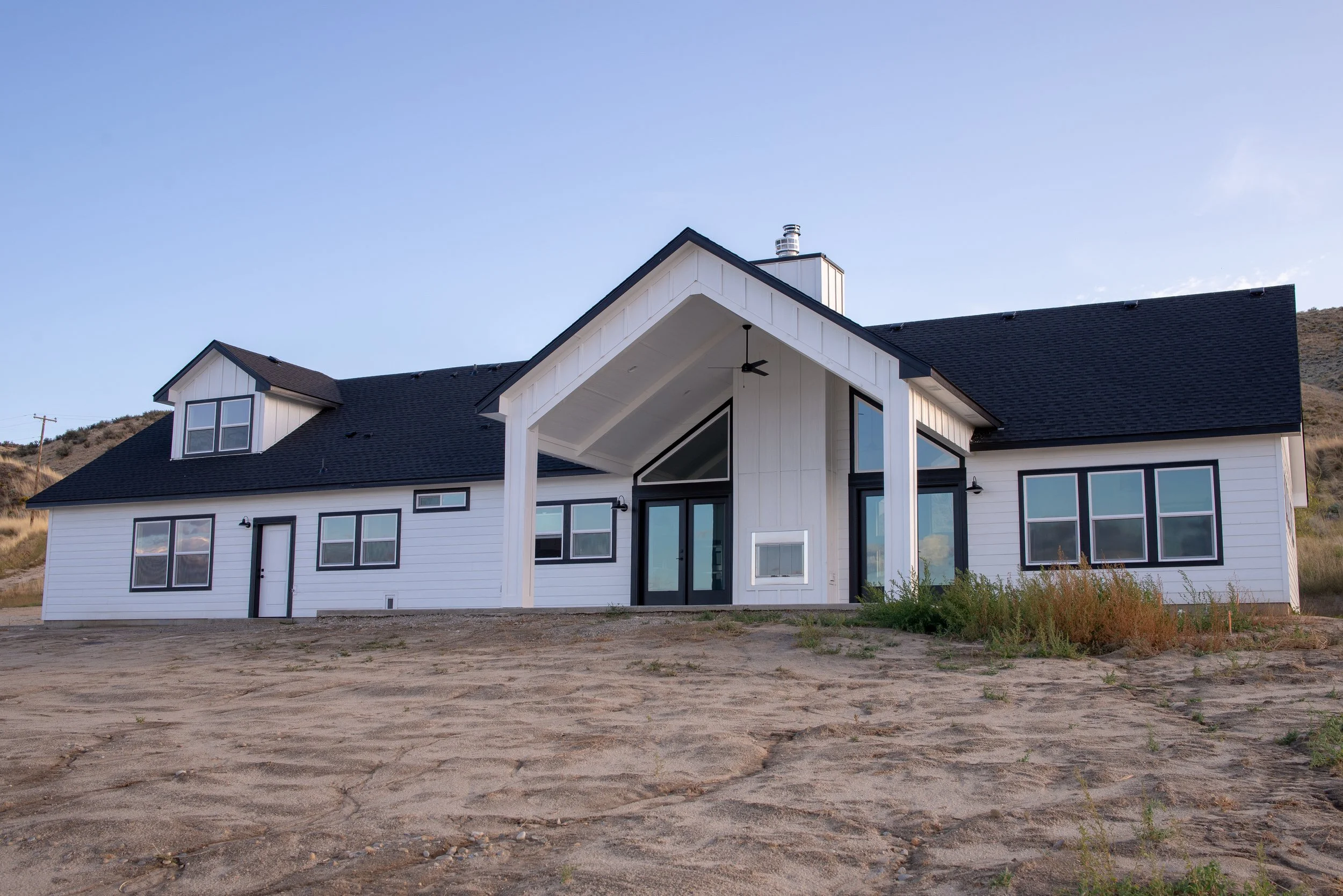A modern white house with black roof shingles and multiple large windows, situated on a lot with bare dirt and some small shrubs, under a clear sky.
