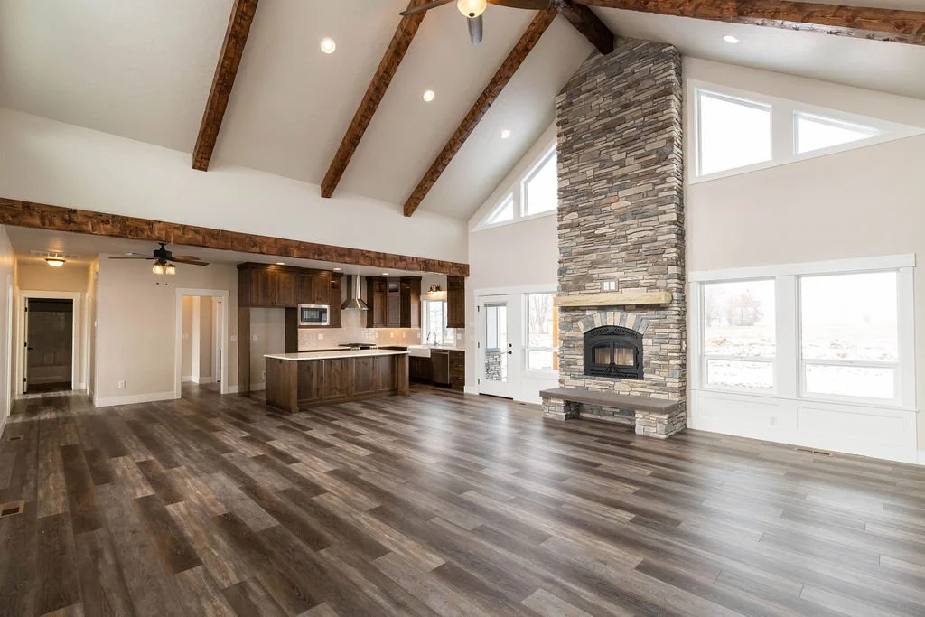 Open-concept living room and kitchen with a vaulted ceiling, wood beams, a stone fireplace, large windows, and hardwood floors.