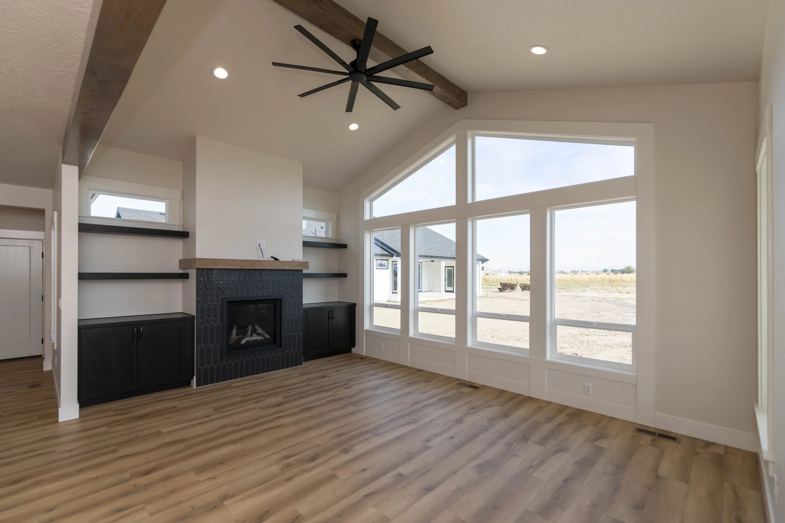 Interior of a modern living room with large windows, a black fireplace with a wooden mantel, wood flooring, and a ceiling fan with multiple blades.