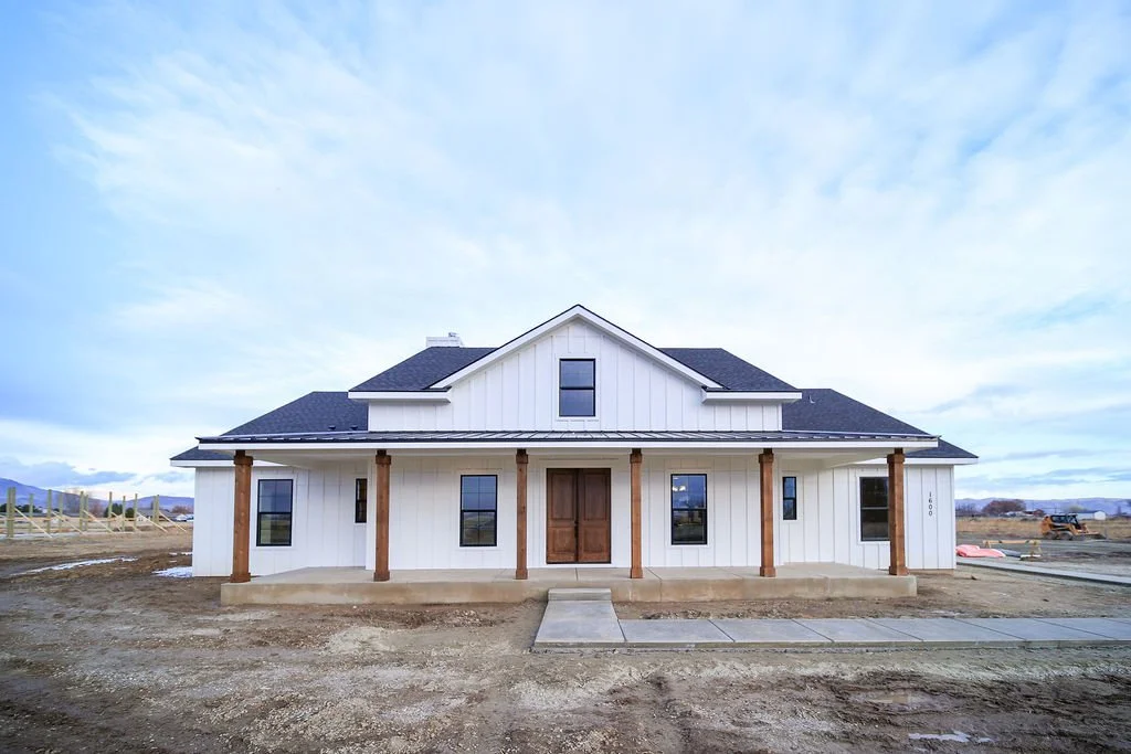 Newly constructed white house with wooden pillars and a front porch, situated in a construction zone with dirt and sidewalk, under a cloudy sky.