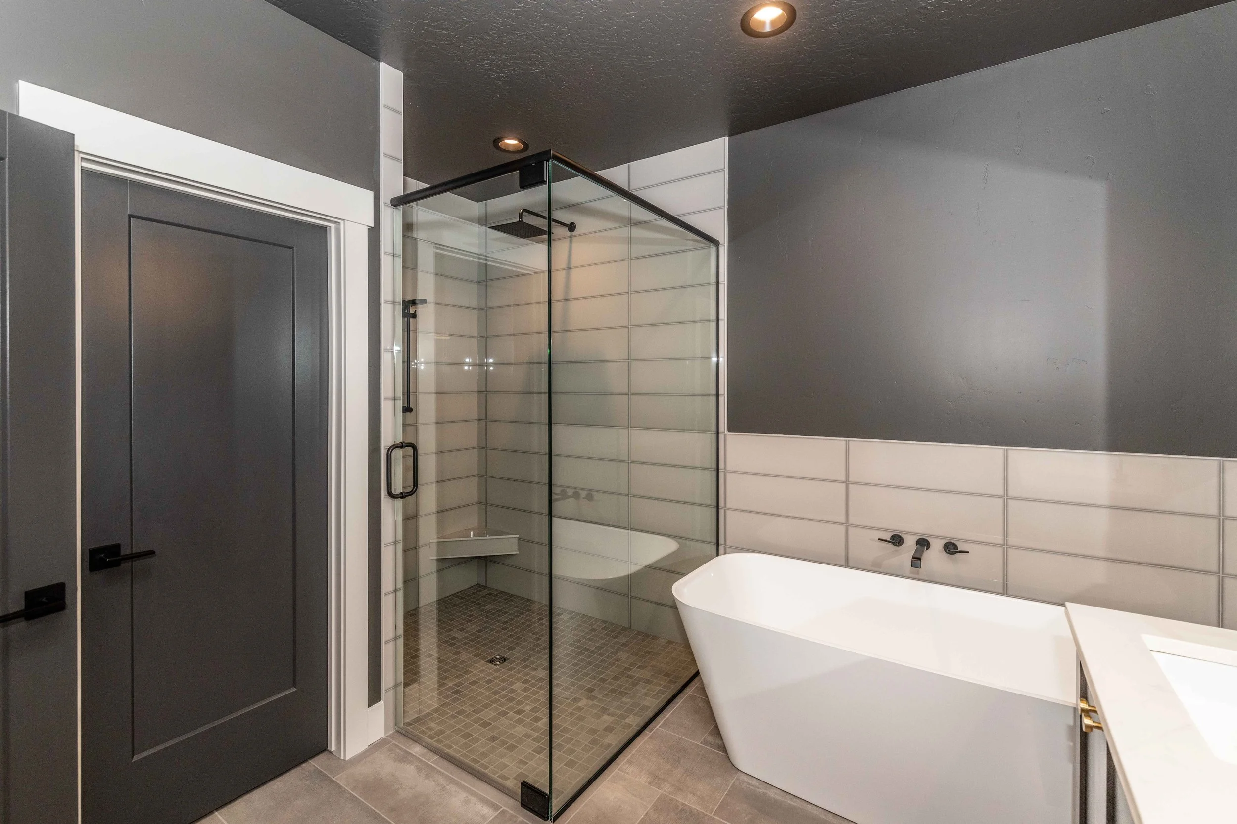 Modern bathroom featuring a bathtub, a glass shower enclosure, dark gray ceiling, white tiled walls, gray door, and gray tiled floor, with recessed ceiling lights.
