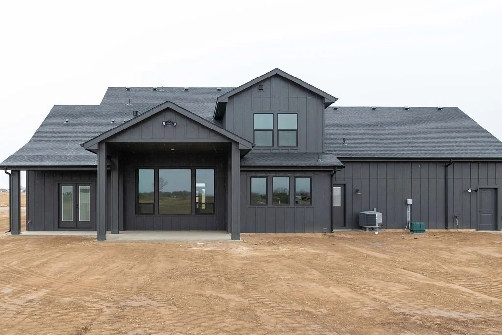 New modern dark gray house with large windows, situated on a dirt lot, under a cloudy sky.