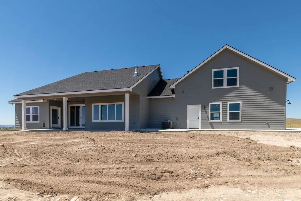 Newly built house with gray siding and multiple windows, with a dirt yard in the foreground, under a clear blue sky
