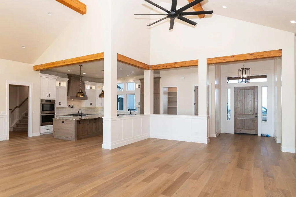 Empty living room with open kitchen, white walls, wooden beams, hardwood floors, and front door with window panels.