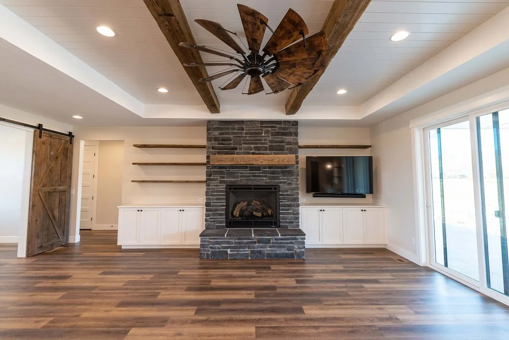 Living room with a stone fireplace, wooden media cabinet, and open shelving. Features a ceiling fan with wooden blades, recessed lighting, and large sliding glass doors.