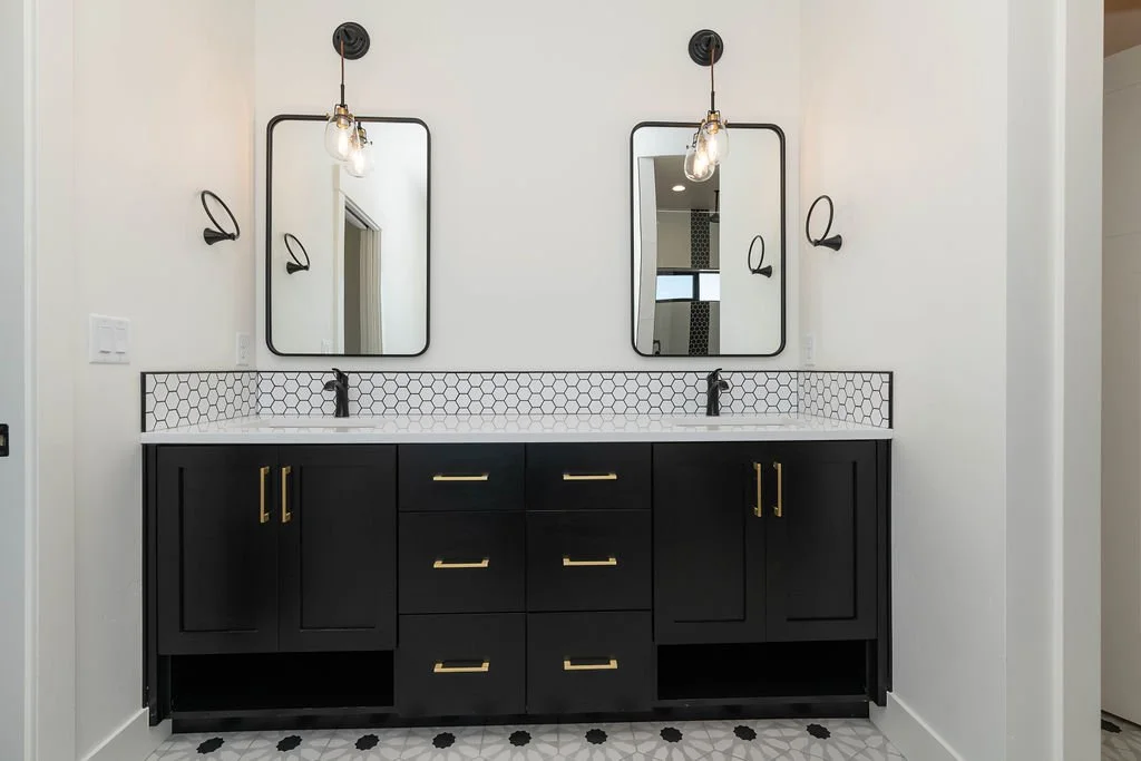 Bathroom vanity with black cabinets, gold handles, two mirrors, black wall sconces, and geometric tile backsplash.
