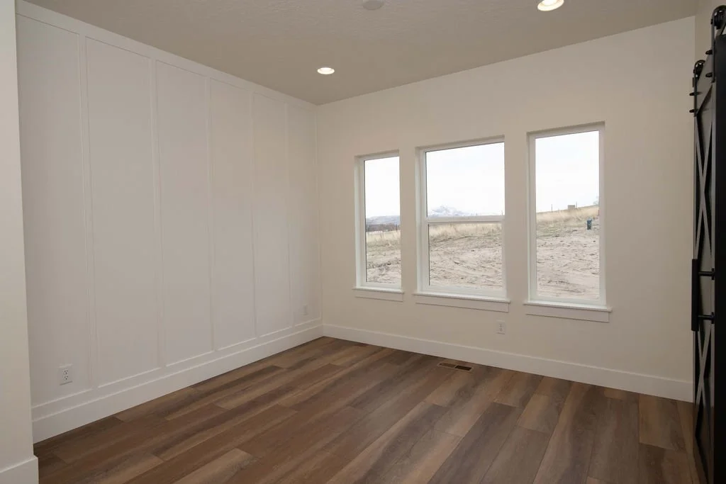 Empty room with three large windows, white walls, wooden floor, and a corner of a black shelving unit.