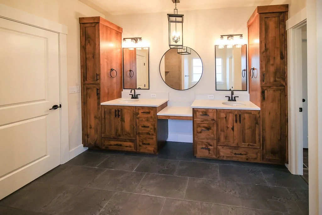 A bathroom with a double vanity featuring wooden cabinets, two mirrors, black faucets, and two light fixtures. A round mirror hangs above the center space, with a hanging light fixture above.