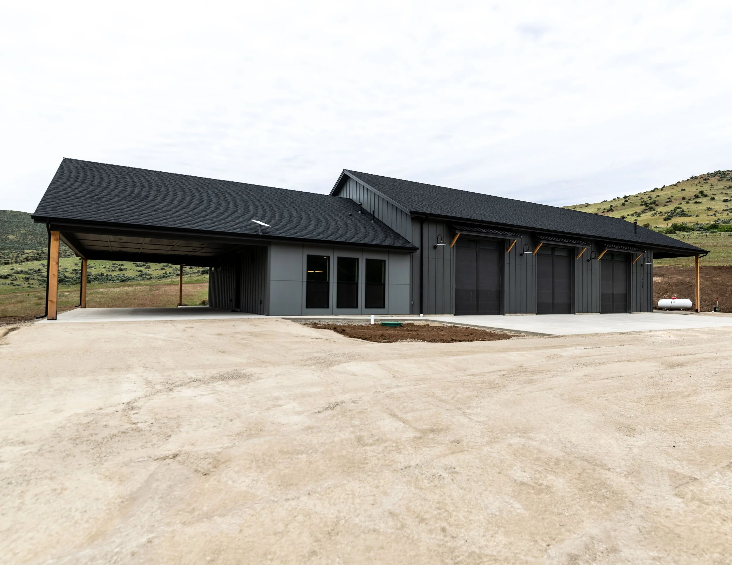 A modern house with a black roof, gray exterior, and large windows situated in a rural landscape with mountains in the background.
