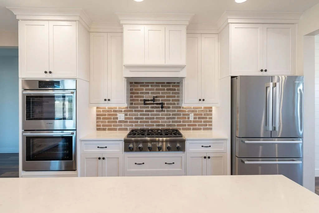 White kitchen cabinets with stainless steel double oven on the left, stove with brick backsplash in the center, and large stainless steel refrigerator on the right.