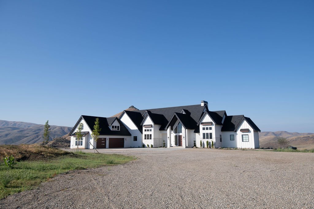 A large white house with a dark roof situated in a rural area with hills in the background, gravel driveway, and minimal landscaping.