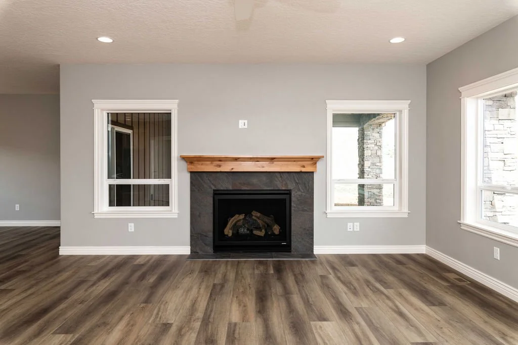 Empty living room with gray walls, hardwood flooring, a fireplace with a wood mantel, and large windows letting in natural light.