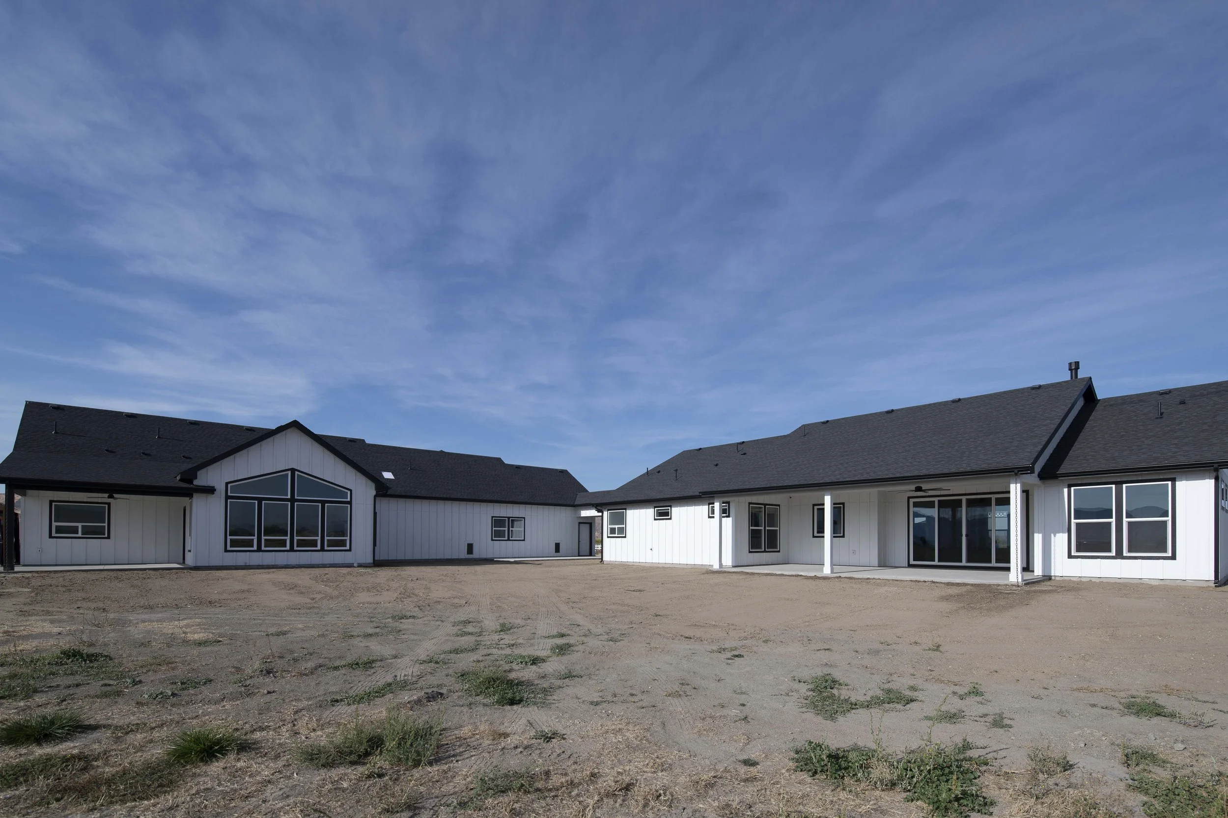 A row of modern white houses with dark roofs under a blue sky with light clouds, a vacant dirt lot in the foreground.