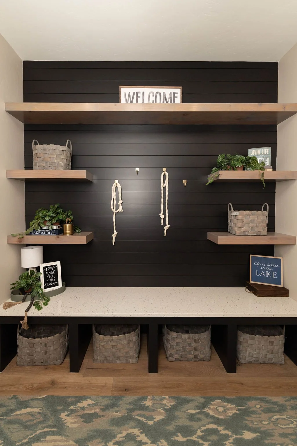 Interior mudroom with black wooden wall, light wood shelves holding decor and plants, and a countertop with storage baskets underneath. Decor includes a 'WELCOME' sign, potted plants, books, and framed signs.
