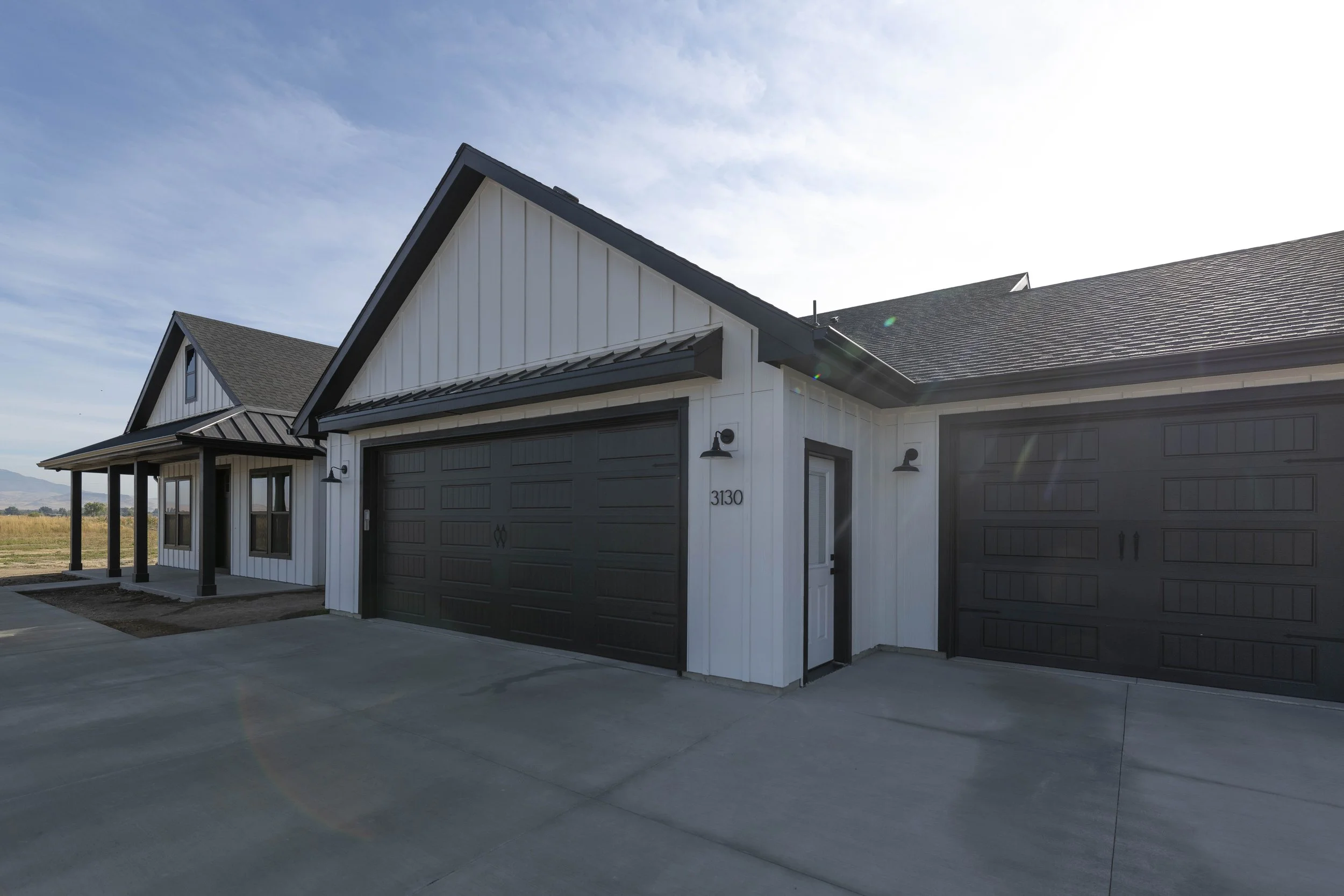 Modern house with black garage doors, white siding, a sloped roof, and outdoor lighting fixtures, located in a rural area with open fields and a clear sky.
