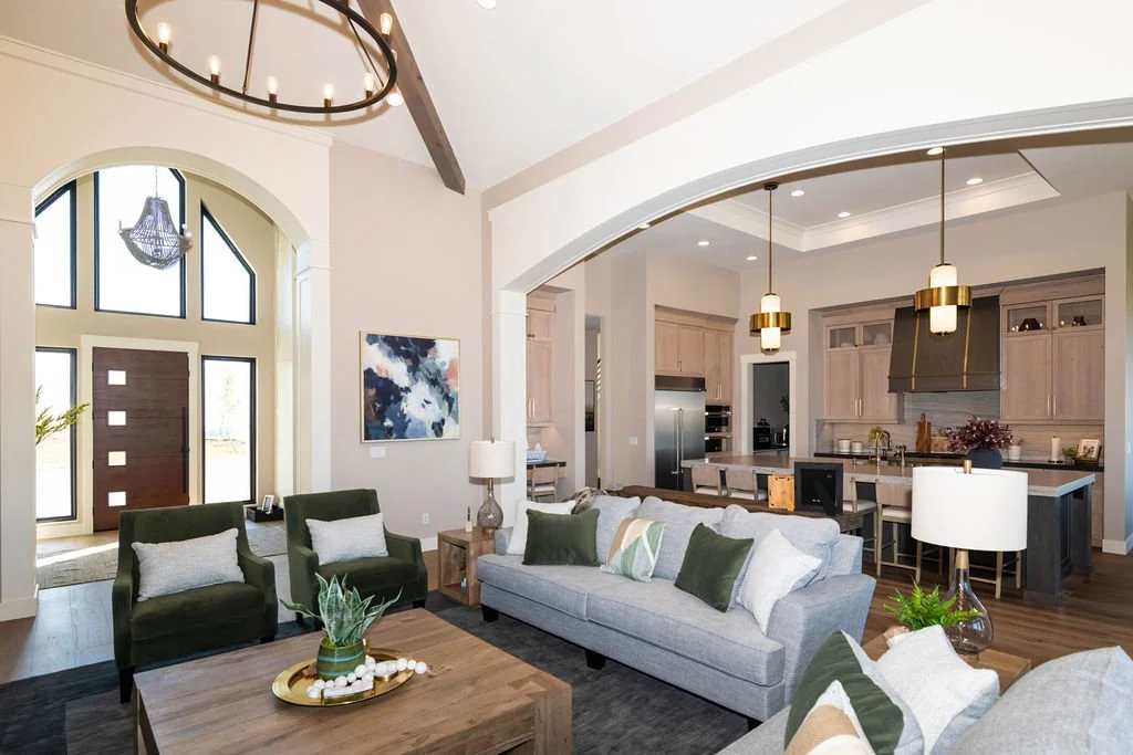Interior of a modern living room and kitchen with high ceilings, large front door, green armchairs, gray sofa, wooden coffee table, dining table, pendant lights, and expansive windows.