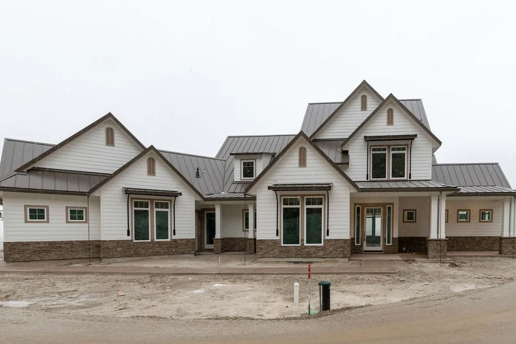 Newly constructed house with white siding, stone accents, and a metal roof, set on a dirt lot.