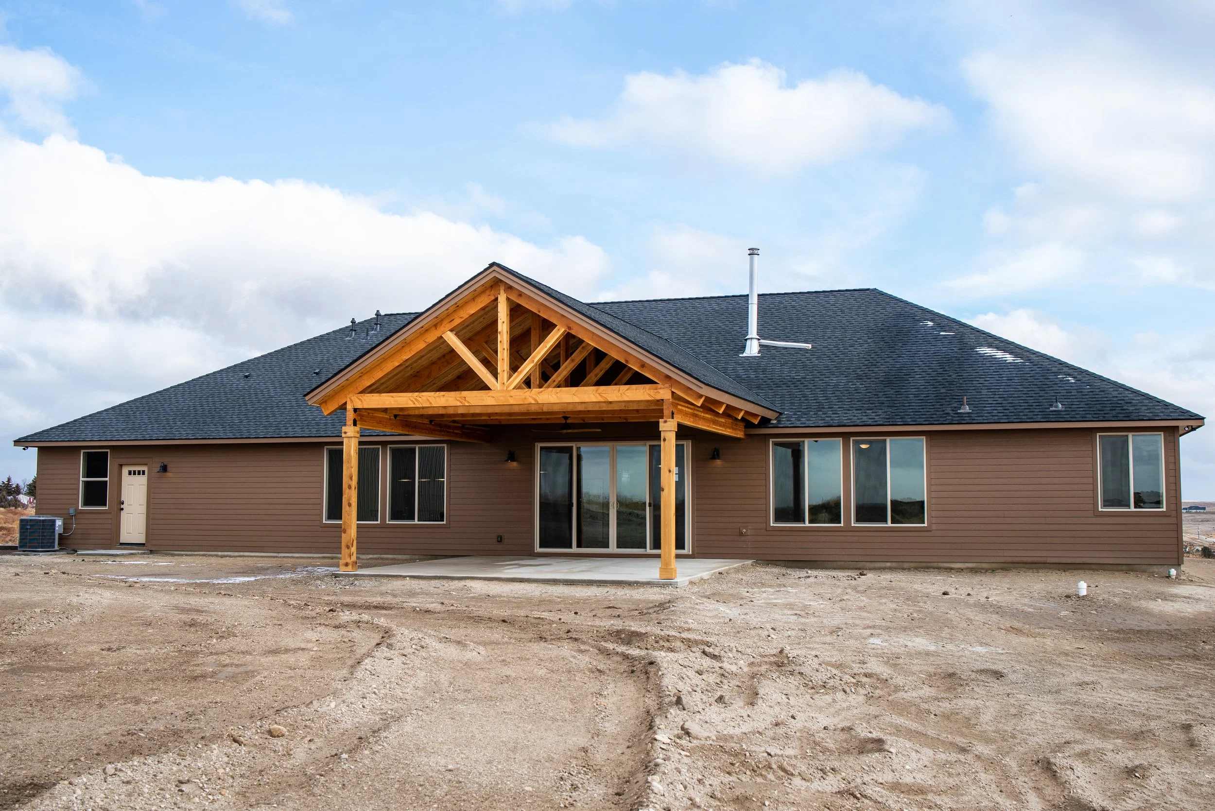 New house with brown siding, large windows, sliding glass door, and a wooden porch structure, under construction or landscaping not yet finished, with an open outdoor area and cloudy sky.