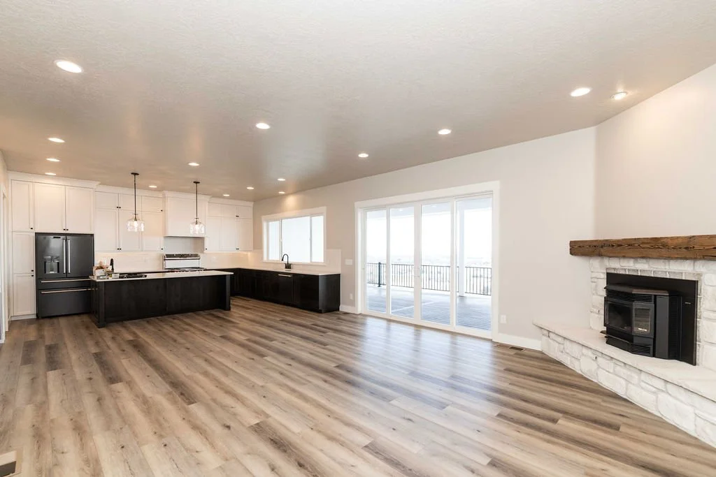 Open-concept living area with a modern kitchen, hardwood floors, and a fireplace with a wooden mantle, leading to a balcony through sliding glass doors.