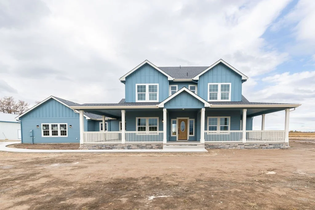 A large two-story blue house with white trim and a wraparound porch, set on a dirt lot under a partly cloudy sky.