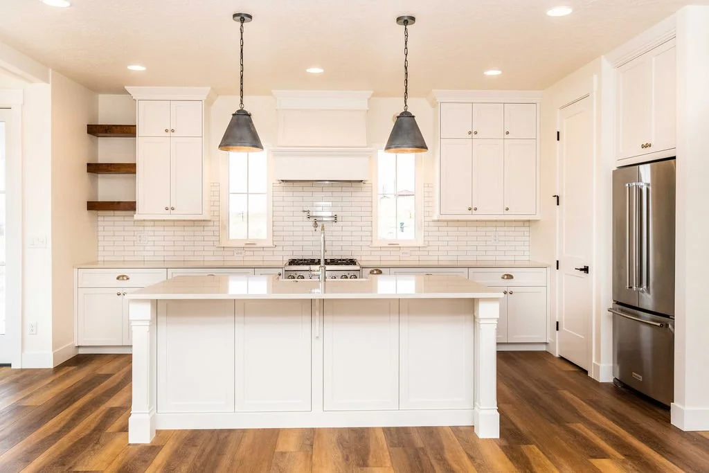 A modern white kitchen with an island, pendant lights, and wooden floor.