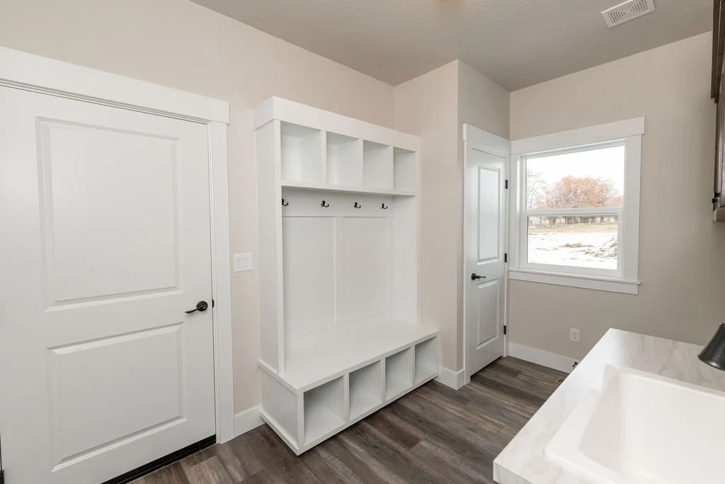 Empty laundry mudroom with white built-in bench, cubbies, hooks, and upper shelves, with a window showing a snowy outdoor scene.
