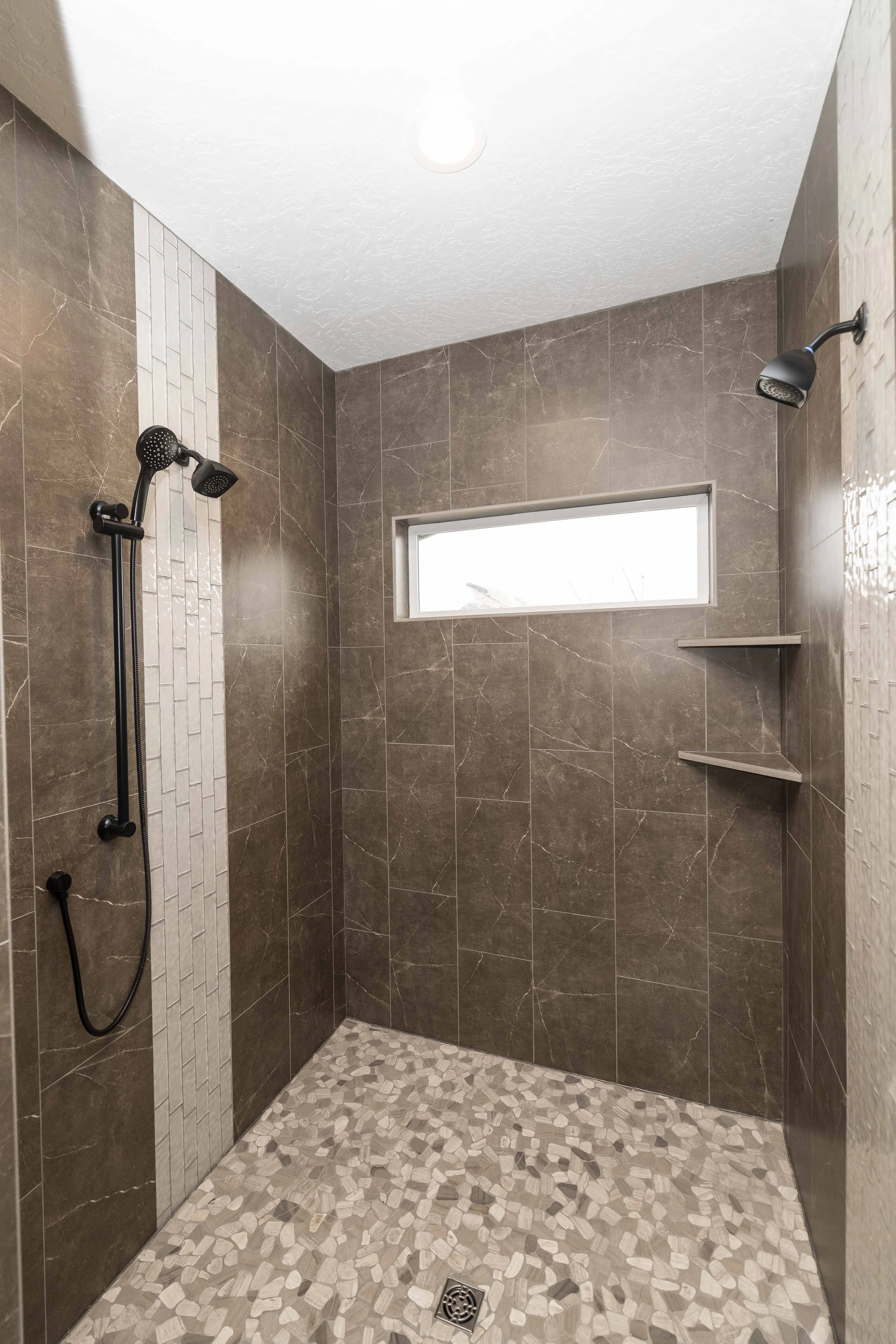 Modern shower with brown marble tile walls, pebble floor, and a small horizontal window. Two black showerheads are mounted on opposite walls, and a metal shelf is in the corner. A ceiling light illuminates the space.