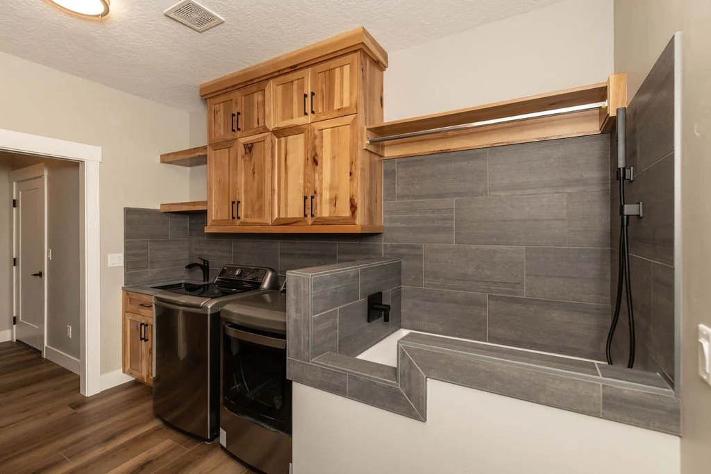 Laundry room with wooden cabinets, gray tiled walls, a washing machine, a dryer, and a laundry sink with a black faucet.