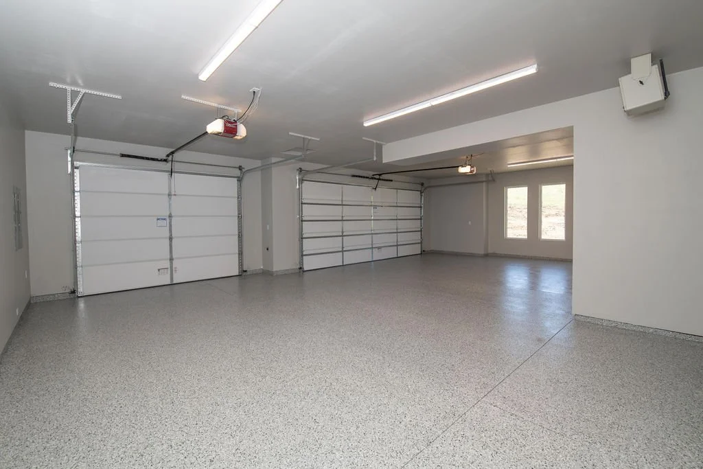 Empty two-car garage with white sectional garage doors, overhead lighting, and a windowed door leading outside.