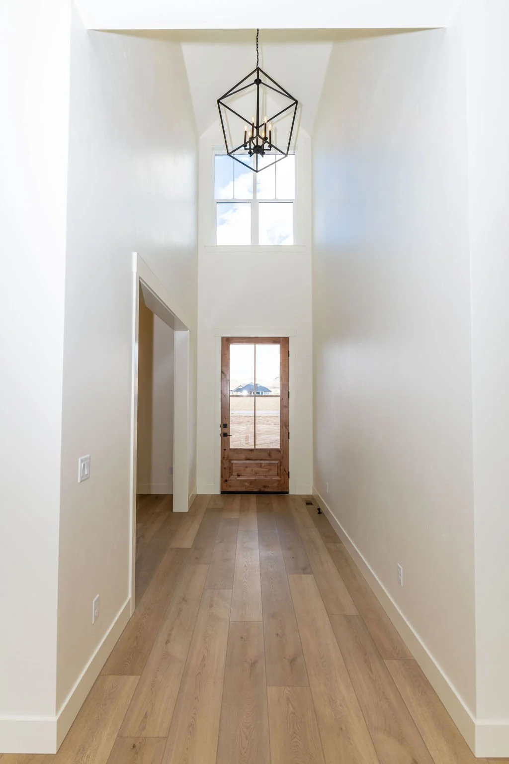 Empty entry hallway with high ceilings, white walls, wooden floor, a hanging geometric chandelier, a window above the wooden door showing an outdoor scene with a house, open doorway on the left, and a wall-mounted switch.