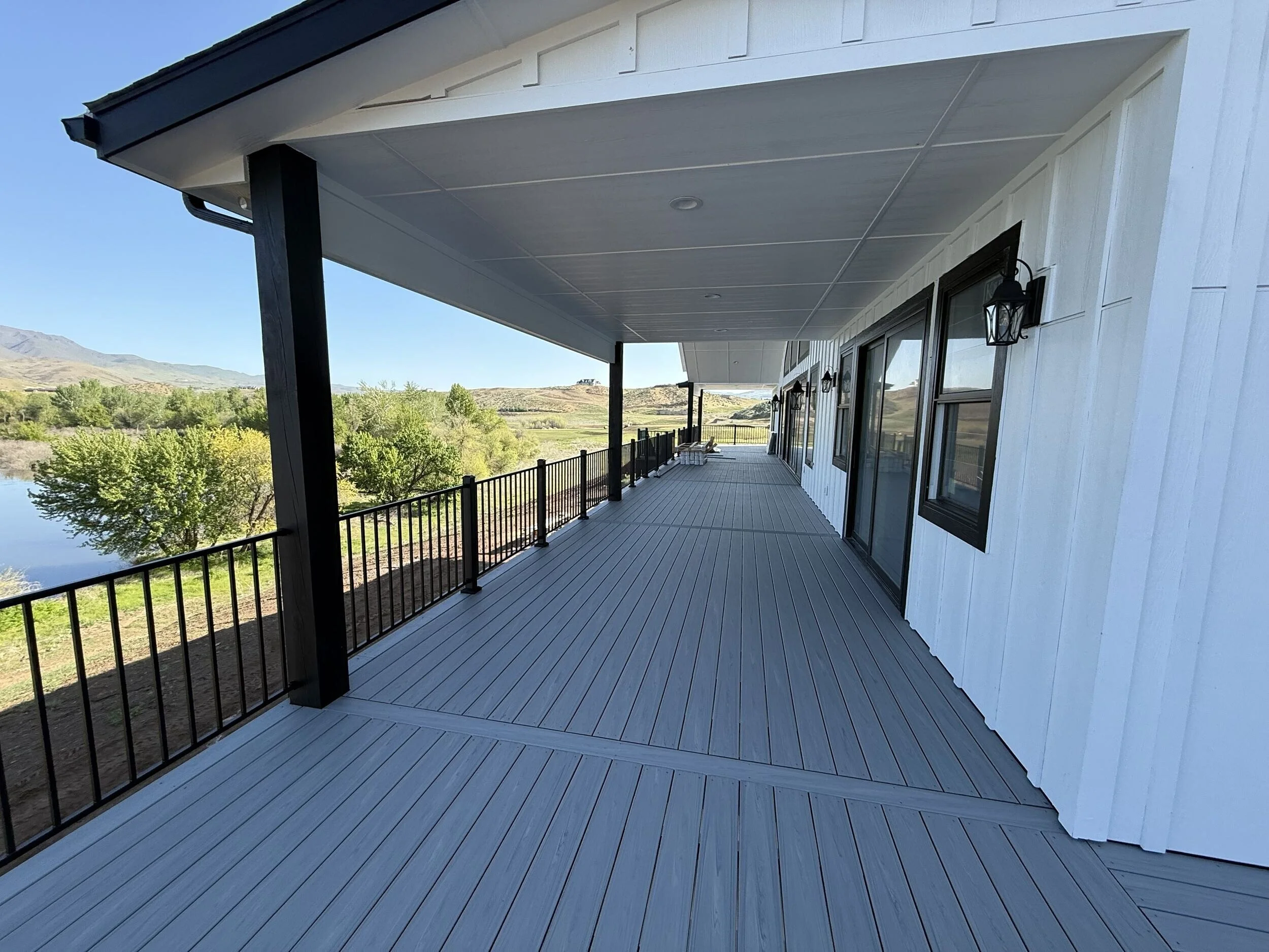 A large outdoor balcony or deck with white wooden flooring and black railings, overlooking a river and green landscape with trees and distant mountains.