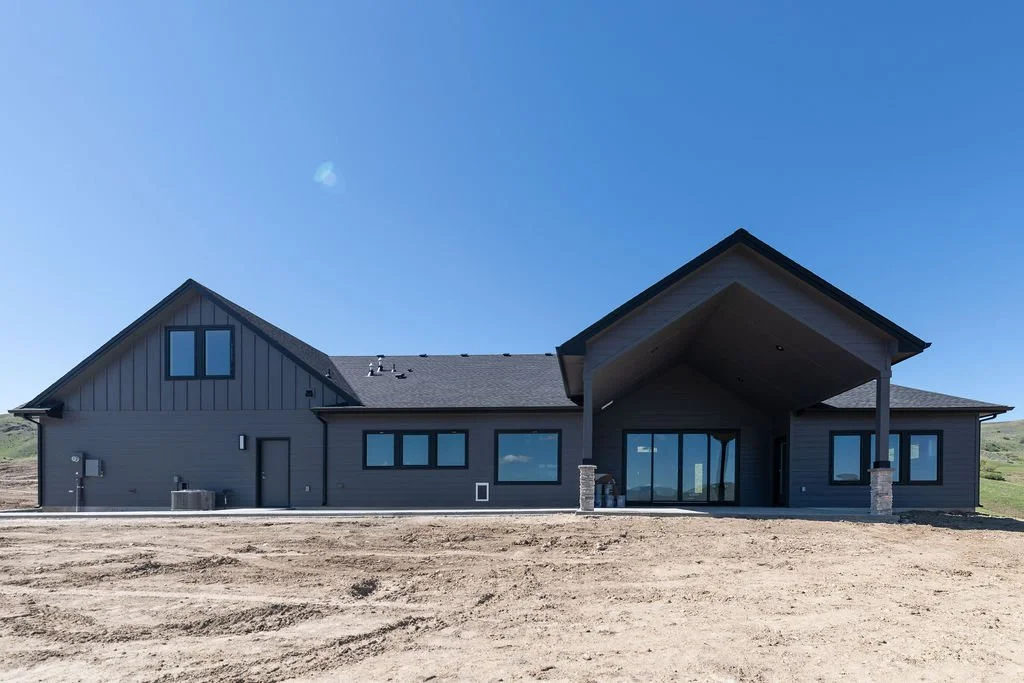 Newly constructed house with dark gray exterior, large glass doors, sloped roof, and a covered porch, situated in a rural area under a clear blue sky.