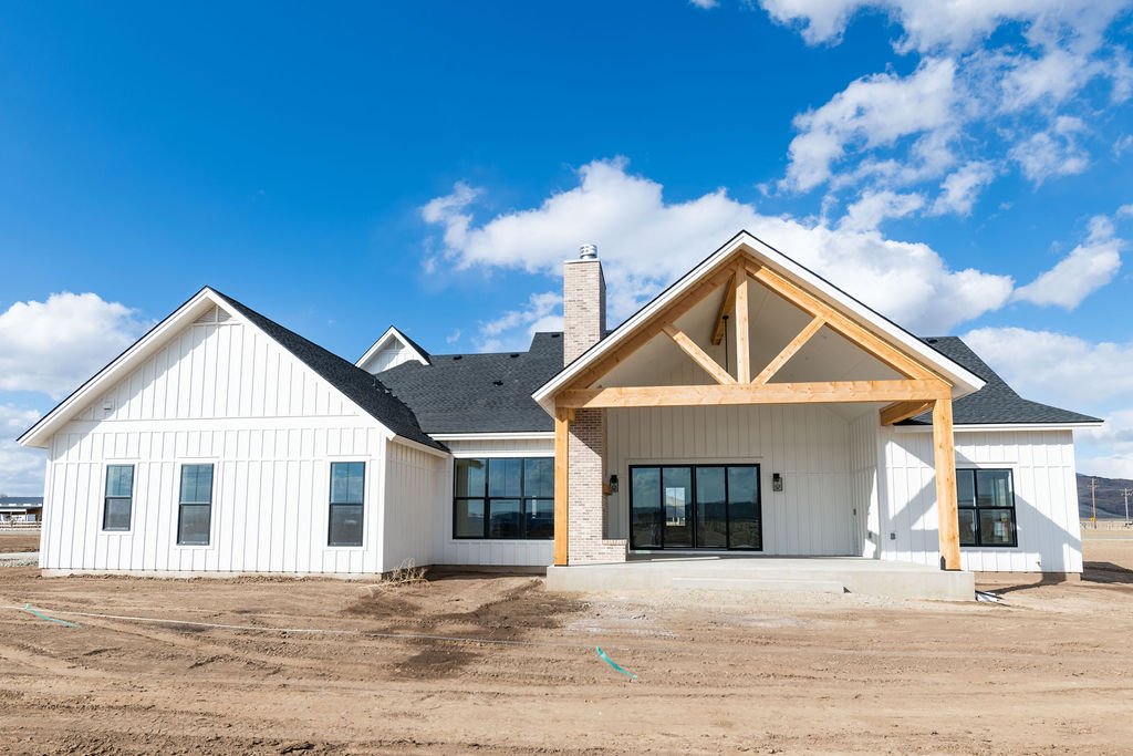 Newly built white house with black roof, large sliding glass door, and exposed wooden beams on the front porch under a partly cloudy blue sky.