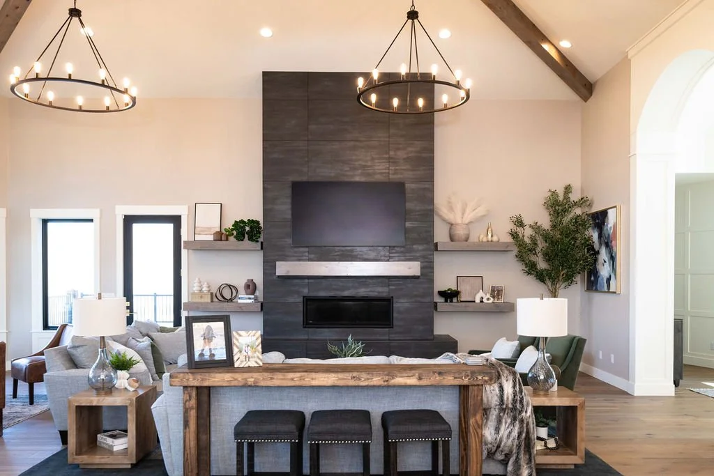 Modern living room with a dark gray fireplace wall, two black ring chandeliers, light-colored walls, and a large window. The room has a sectional sofa, side tables with lamps, and decorative shelves with various ornaments and plants.