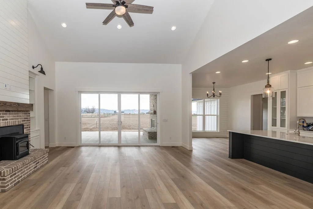 Empty living room with wooden flooring, a brick fireplace, and large sliding glass doors leading outside. Open kitchen area with white cabinets, island with black base, and pendant lighting.