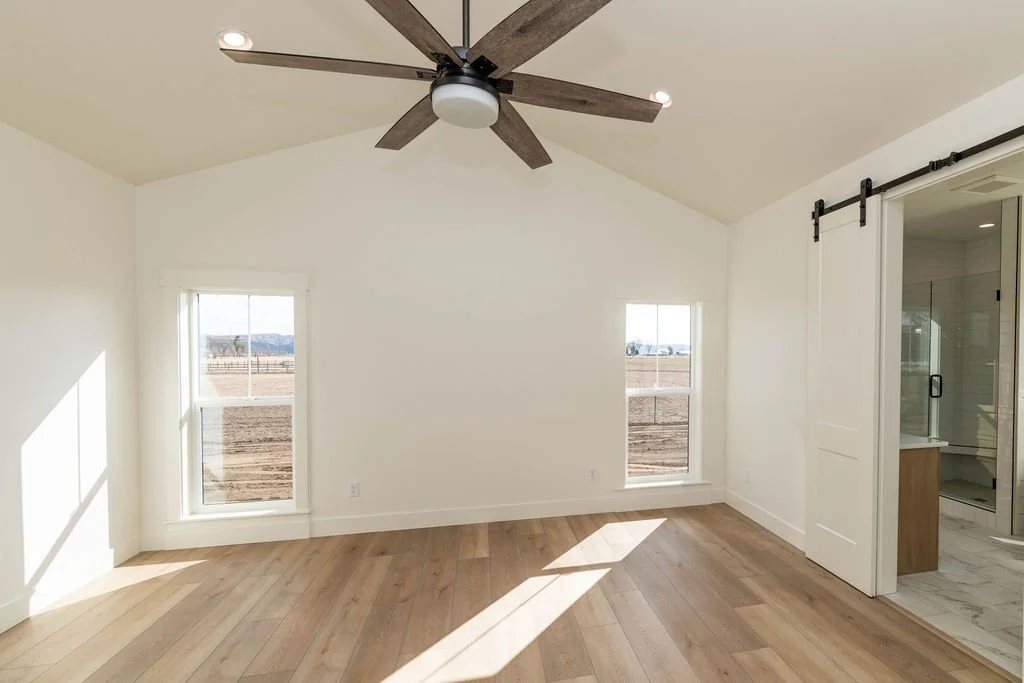 Empty room with white walls, two windows with white frames, a ceiling fan with wooden blades, and a sliding barn door leading to a bathroom with a shower.