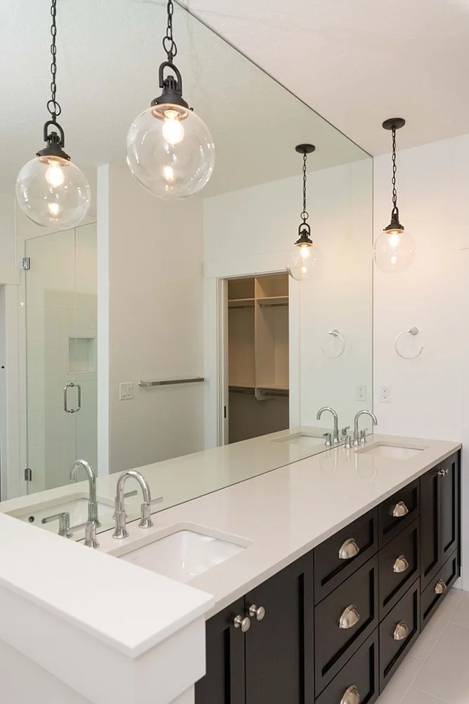 Modern bathroom vanity with a large mirror, three light fixtures with glass globes, double sinks with chrome faucets, and dark cabinetry with gold knobs. A glass-enclosed shower is visible in the background, and a small open closet with shelves is ne