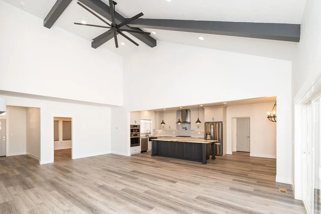 Open-concept living area with high white walls, light wood flooring, a large black ceiling fan, and a modern kitchen with black cabinets, stainless steel appliances, and pendant lighting.
