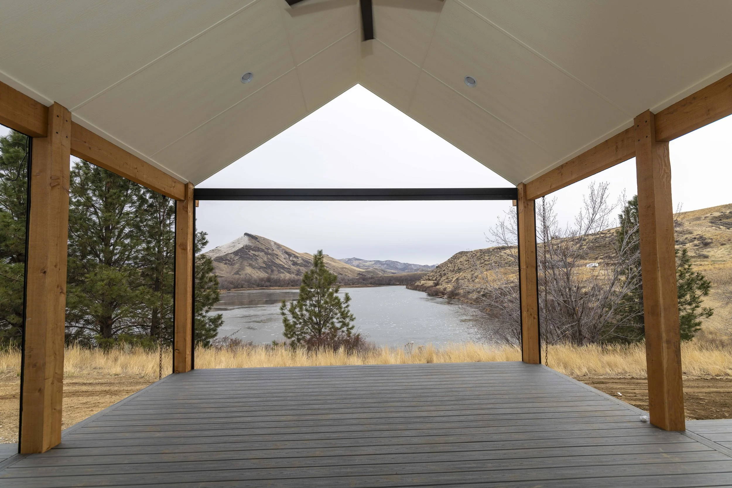 A wooden deck with a roof overlooking a river, trees, and mountains in the distance.