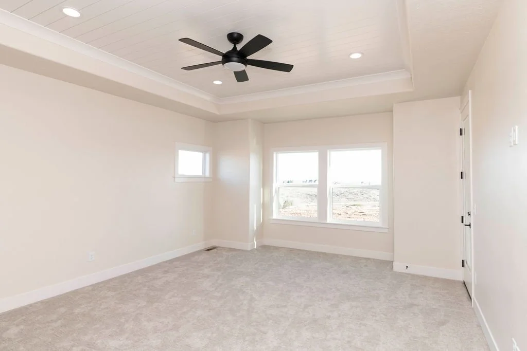 Empty bedroom with beige carpet, white walls, a black ceiling fan, and three windows allowing natural light.