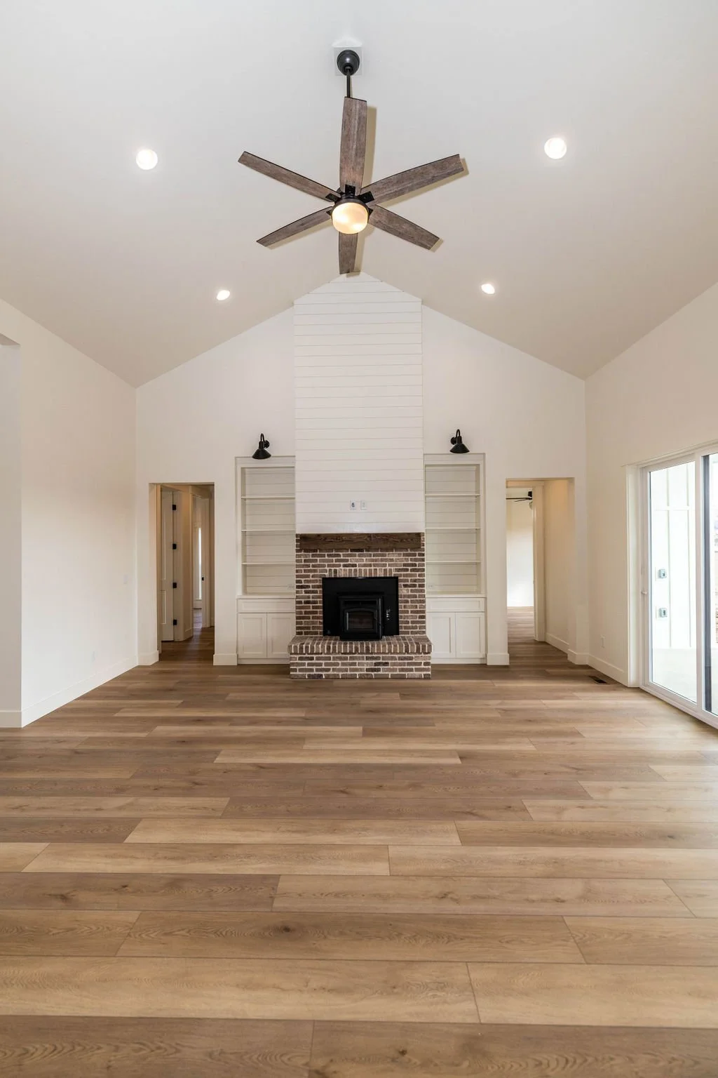 Empty living room with a brick fireplace, white built-in shelves, a ceiling fan, and large windows with sliding glass doors.