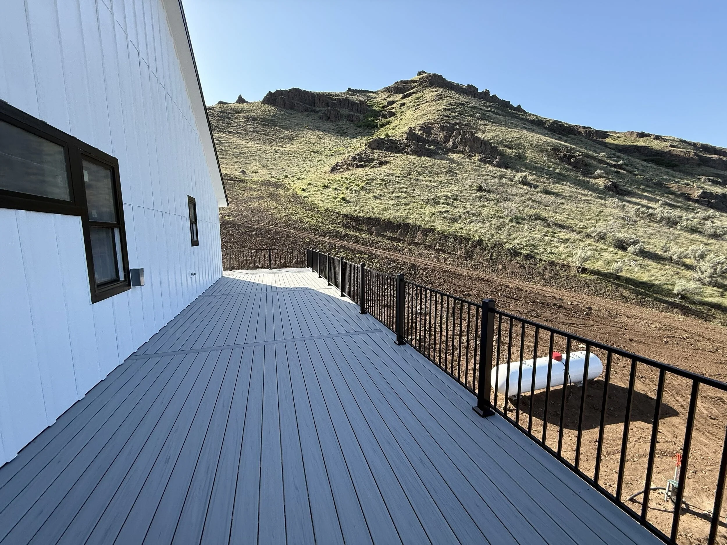 View from a deck of a white house with black-framed windows, overlooking a hillside with sparse grass and rocky formations under a clear blue sky.