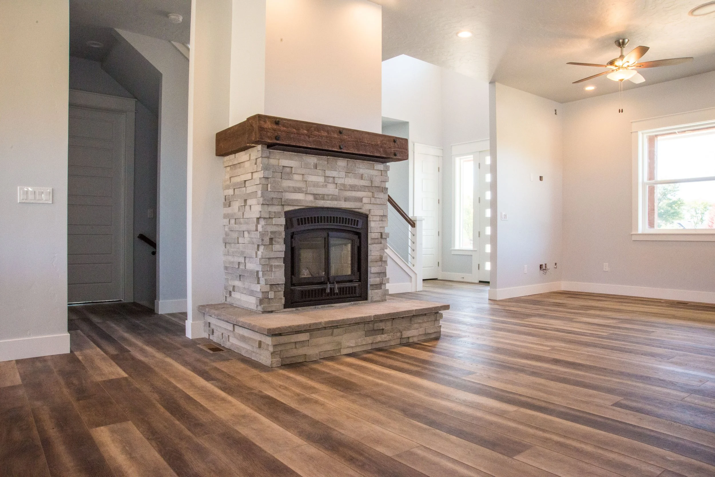 Empty living room with a brick fireplace, hardwood floors, white walls, large windows, a ceiling fan, and a doorway.