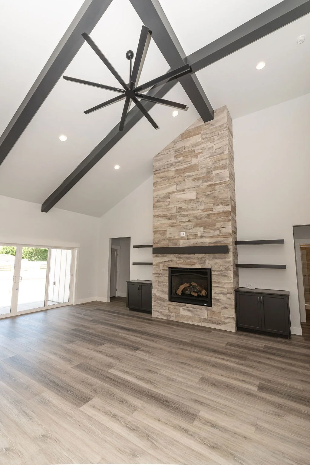 Modern living room with a high ceiling, wood floors, a stone fireplace with a chimney, black cabinetry on either side, a black ceiling fan, and sliding glass doors leading outside.