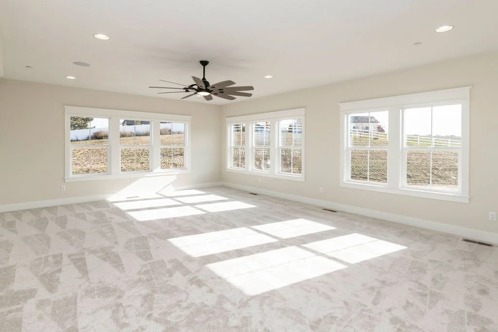 Empty living room with large windows, white walls, beige carpet, and a ceiling fan.