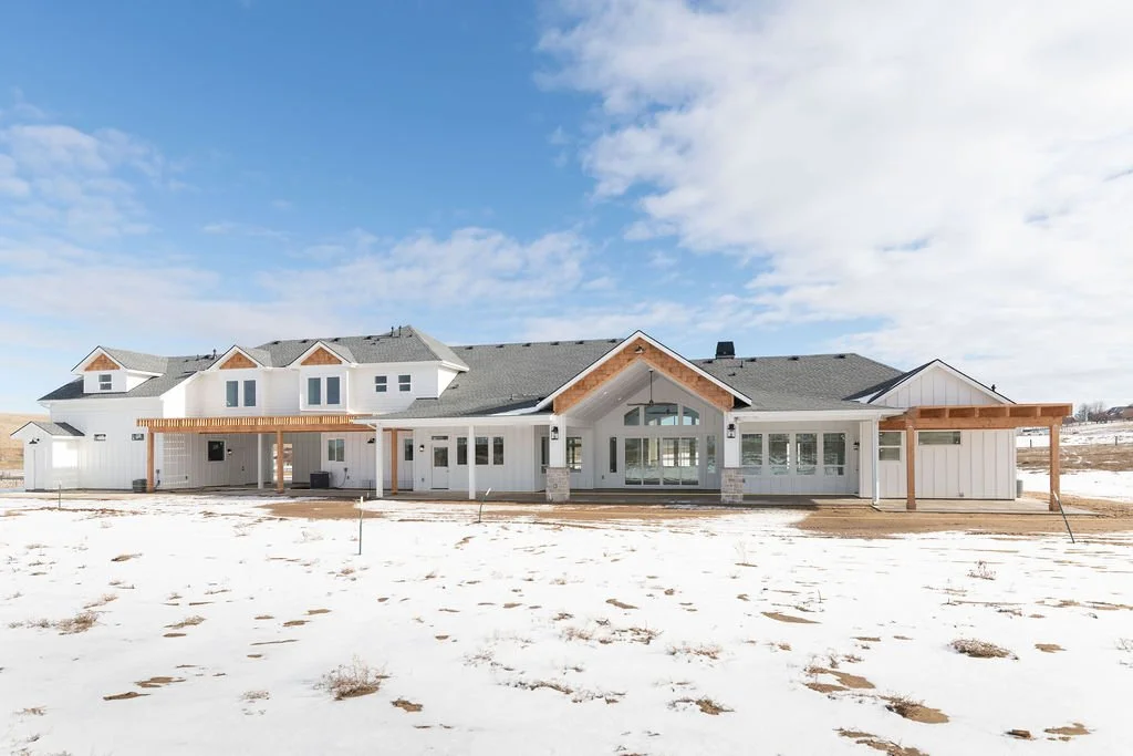 Modern two-story house with white siding, black roof, and wooden accents, surrounded by snow and under a partly cloudy sky.