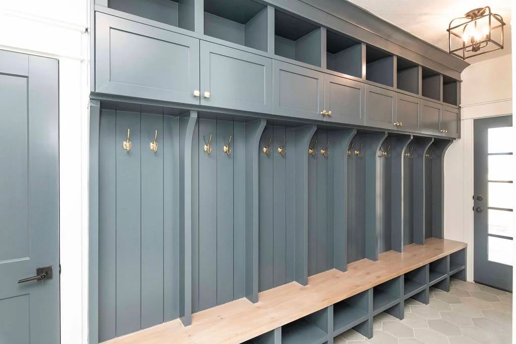 Blue mudroom with built-in cubbies, gold hooks, and upper cabinets, next to a door with glass panels.