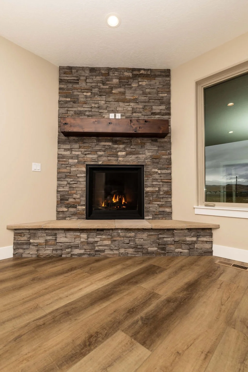 Living room corner with stone fireplace, wooden mantle, beige walls, and large window showing outdoor landscape.