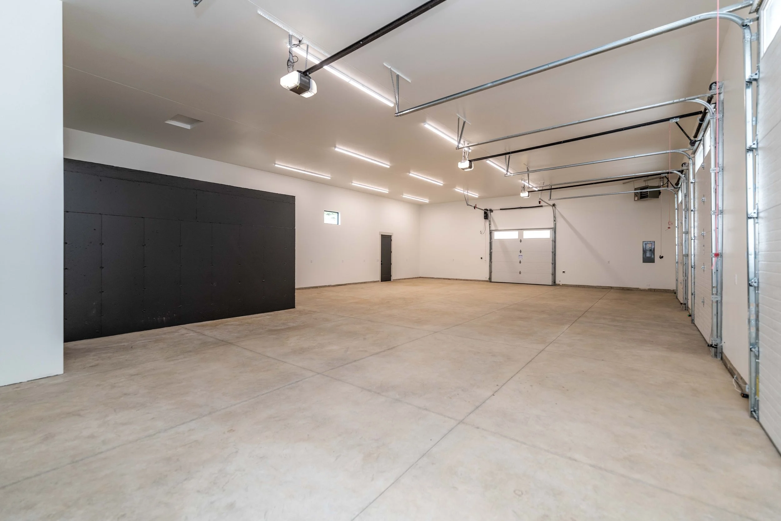 Empty garage with concrete floor, white walls, a black partition on the left, and a garage door with windows on the right. Ceiling has strip lighting and several garage door openers and rails.