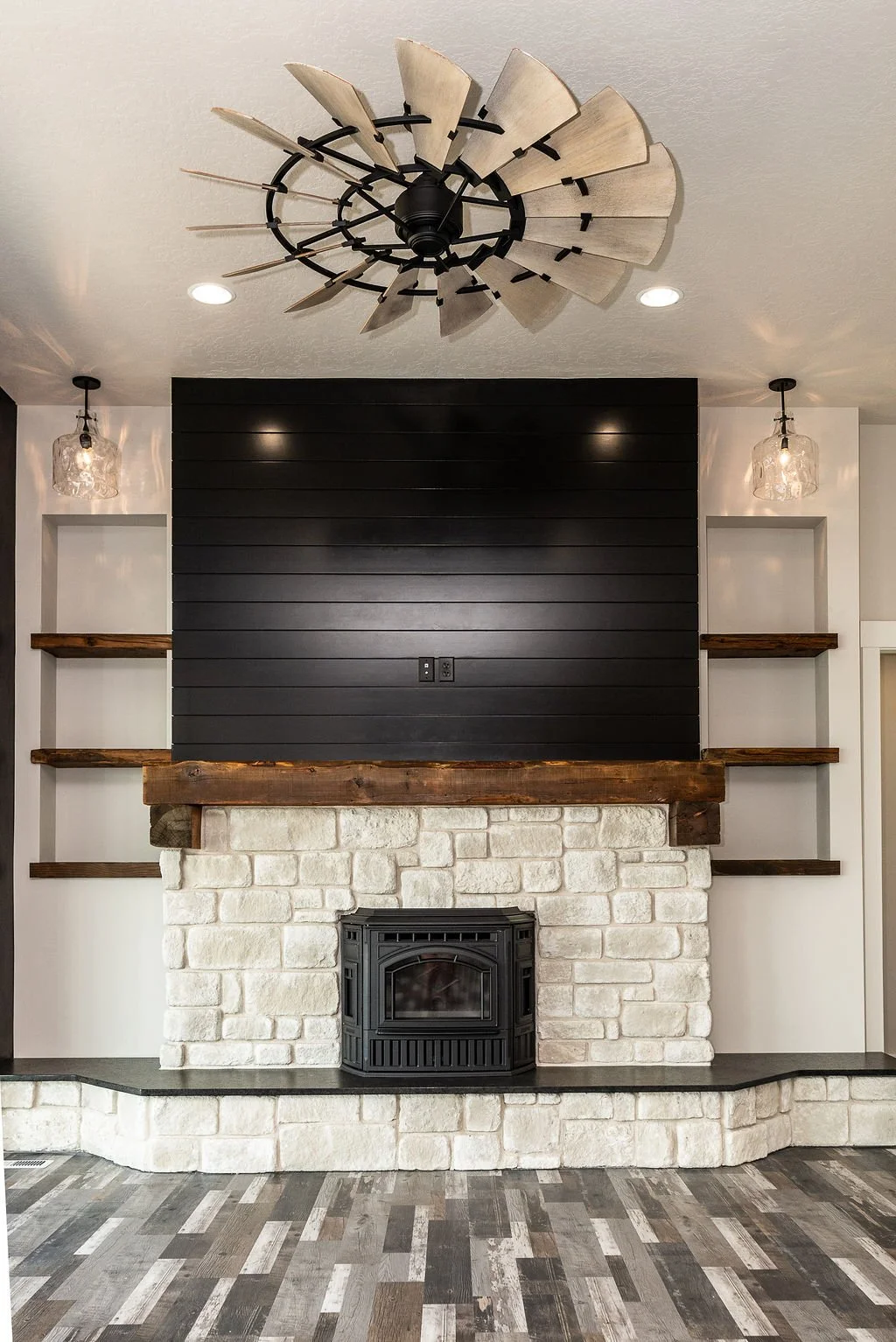 A living room fireplace with a black wood stove, white stone surround, a dark wood mantel, black shiplap on the wall above the mantel, and built-in shelves with wooden accents. A modern ceiling fan with wood blades is above, with two light fixtures o